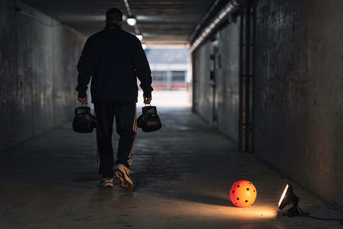 Pale dawn light in a concrete stadium service tunnel as a handler seen from behind carries boxing gloves past a torch left burning on the ground beside a still.