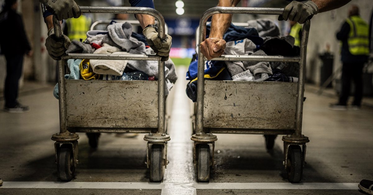 Close-up in a floodlit stadium tunnel of two pairs of gloved hands gripping the handles of loaded metal laundry trolleys on painted concrete, taped start line.