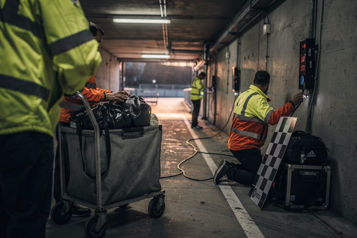 Wide shot down a long stadium tunnel at dusk, part-cropped groundstaff in high-vis jackets fixing electronic timing gates to the walls while a checkered flag.