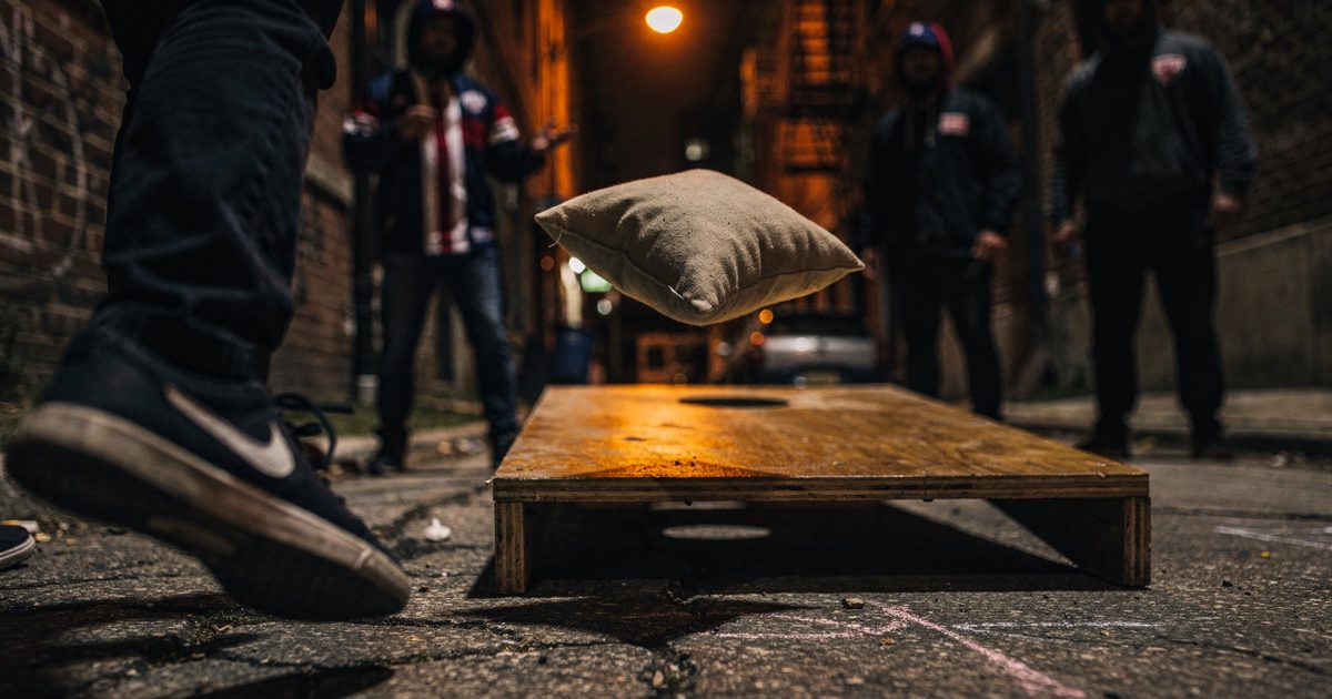 Close detail in a Chicago alley at night under one orange sodium streetlight, a canvas beanbag frozen mid-flight inches above a plywood cornhole board, a.