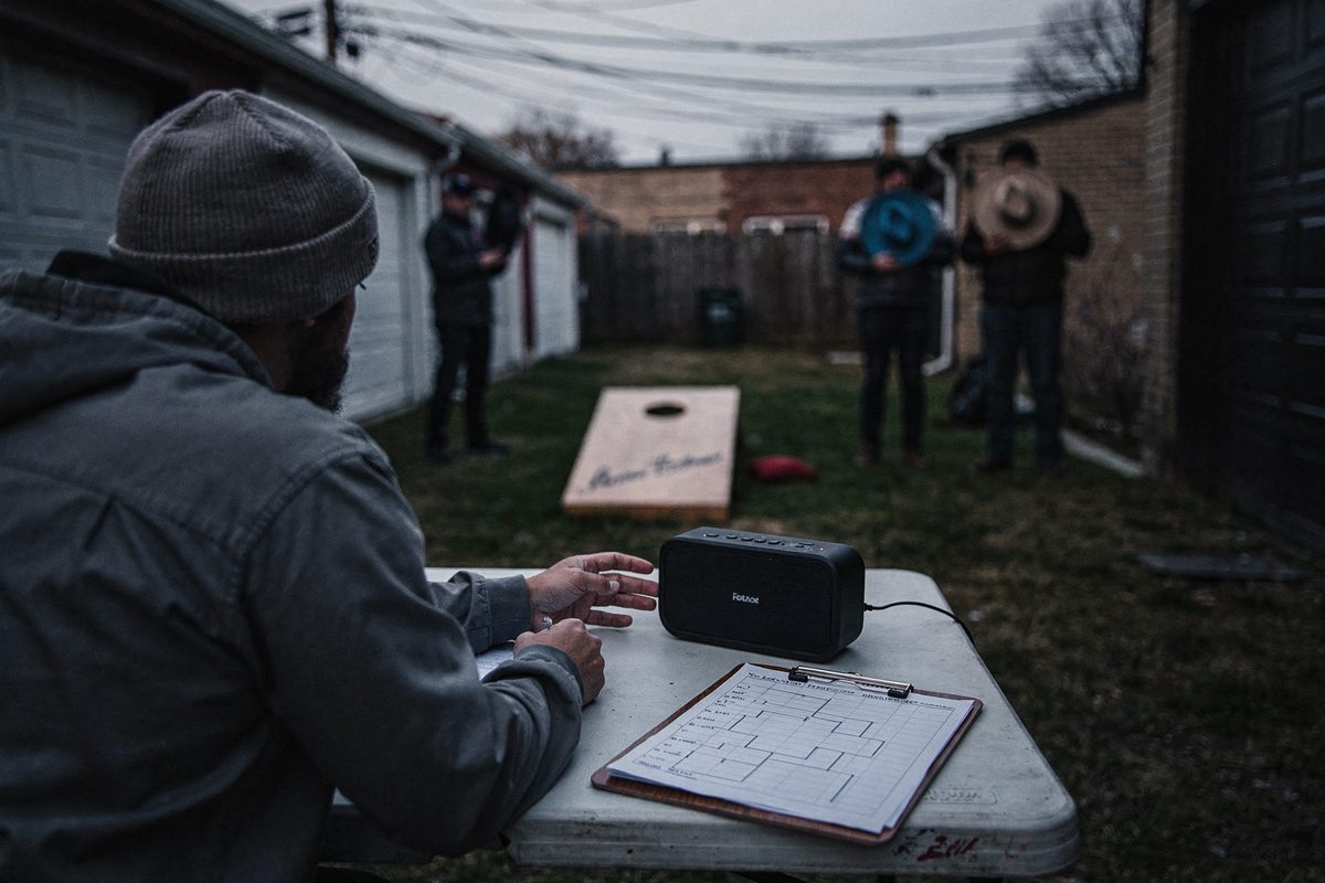 Medium shot from behind a folding card table in a garage-lined alley at dusk, a volunteer scorekeeper in a knit cap reaching for a paused portable speaker, a.