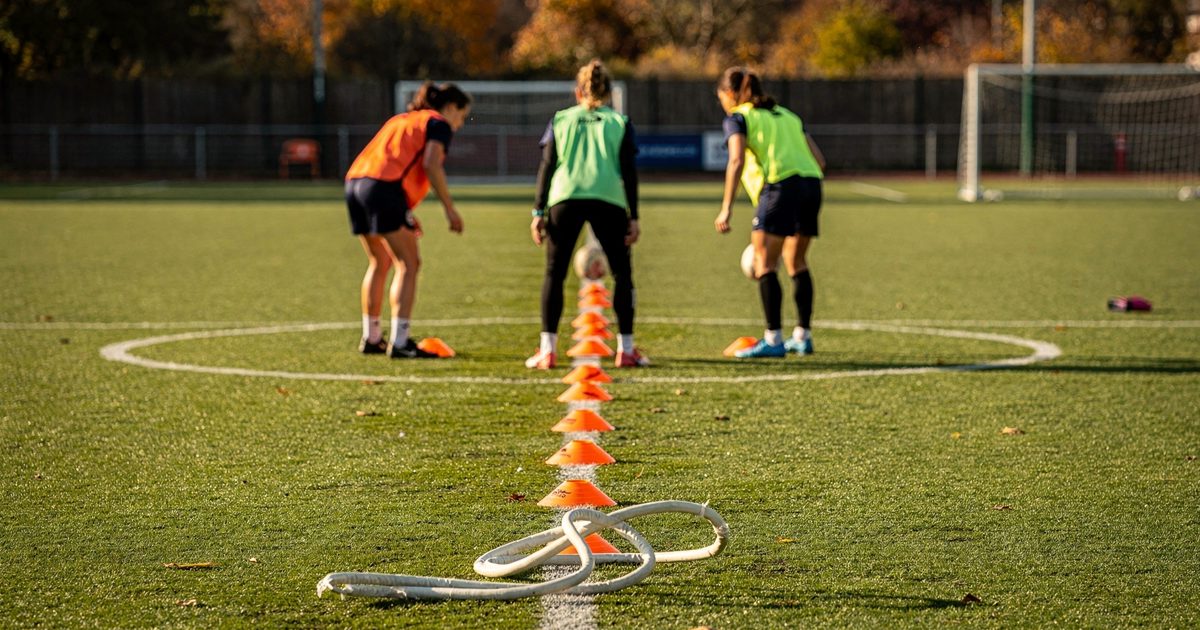 Wide shot in sharp early-autumn sunlight across a grass football training pitch, three players seen from behind in training bibs placing orange cones through.