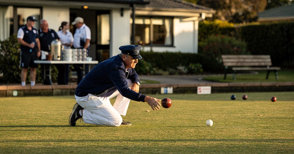 Low angle across a flat suburban bowling green in bright early-evening sun, a player in a navy tracksuit top and white flannels crouched mid-delivery with his.