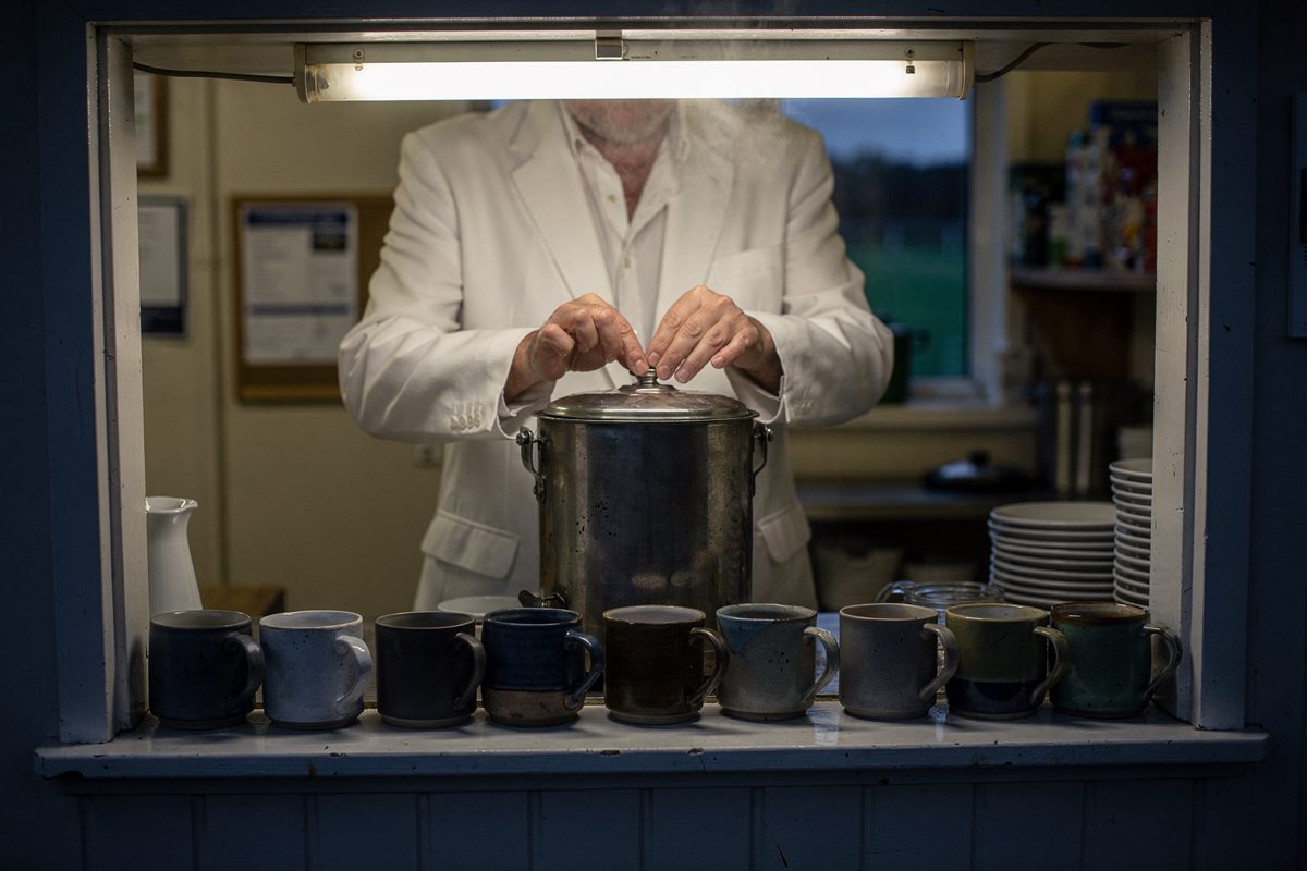 Medium shot through the serving hatch of a small bowls clubhouse kitchen at dusk under a fluorescent tube, a steward in a white blazer cropped at the shoulders.