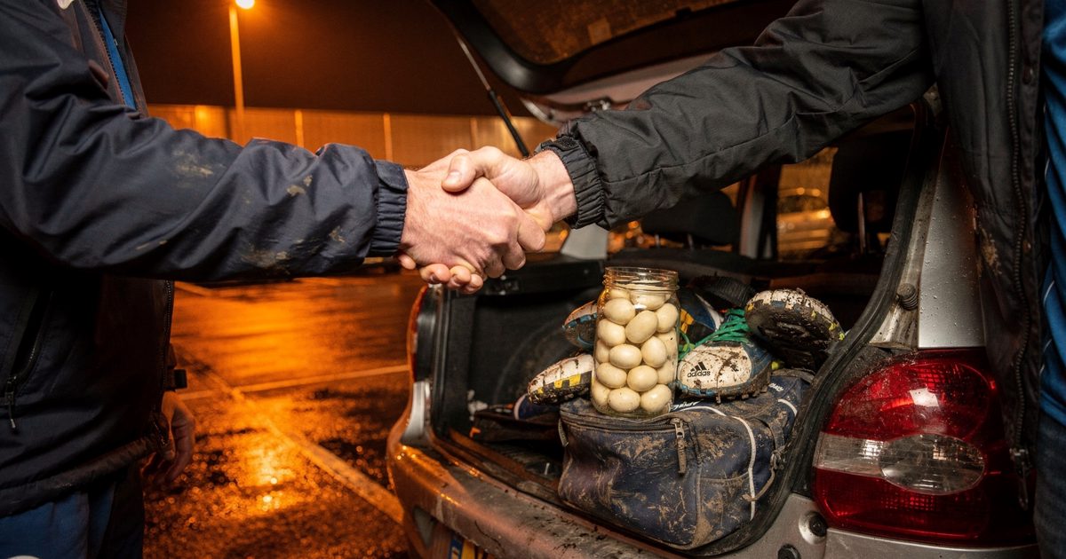 Tight close-up at night in a leisure centre car park under a single orange sodium floodlight, two hands mid-handshake over the open boot of a small hatchback.