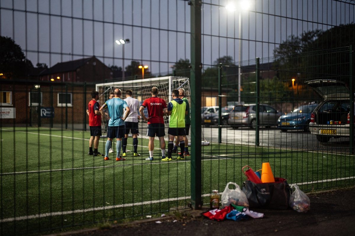 Wide shot through the chain-link fence of an outdoor artificial five-a-side pitch on a clear Friday evening, floodlights blazing white, a handful of amateur.