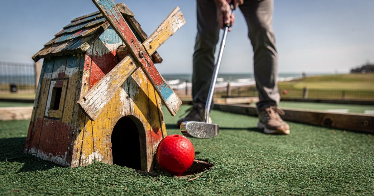 Ground-level shot at a seaside crazy golf course in bright Sunday-morning sun, a red golf ball resting at the mouth of a painted wooden windmill while a.