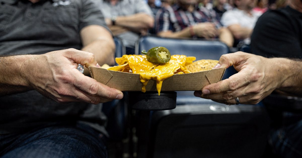 Tight low-angle close-up of two men's hands gripping opposite edges of a cardboard nacho tray balanced on a stadium armrest, congealed cheese and a single.