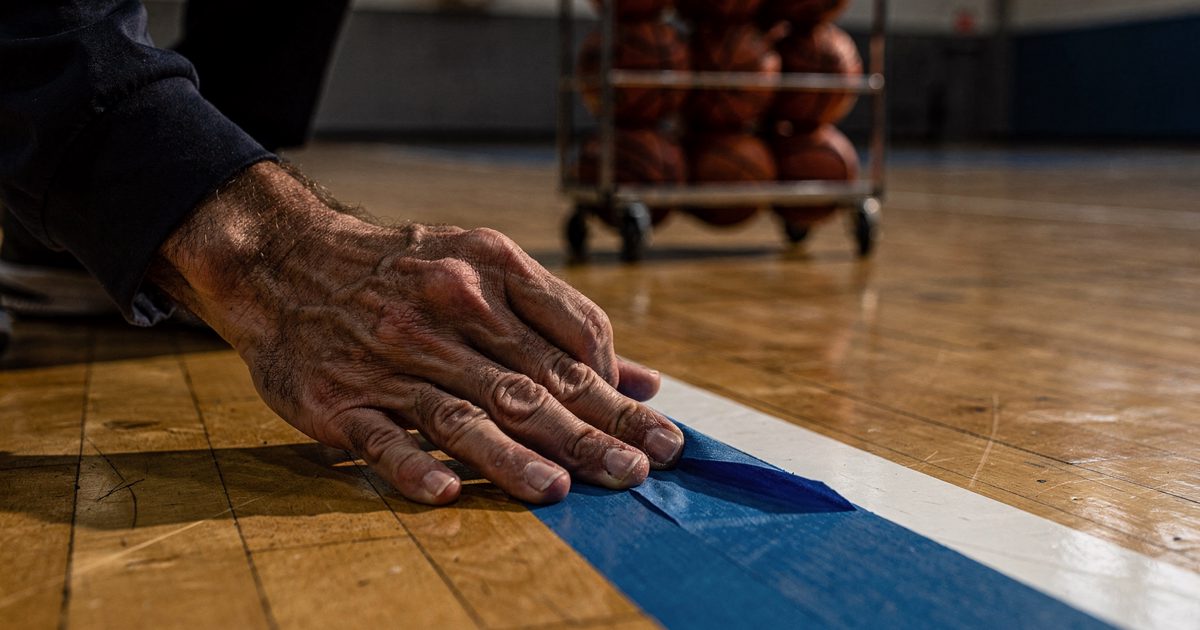 Close-up in low morning window light of a maintenance worker's hands pressing fresh blue painter's tape onto a hardwood free-throw line, a rack of basketballs.