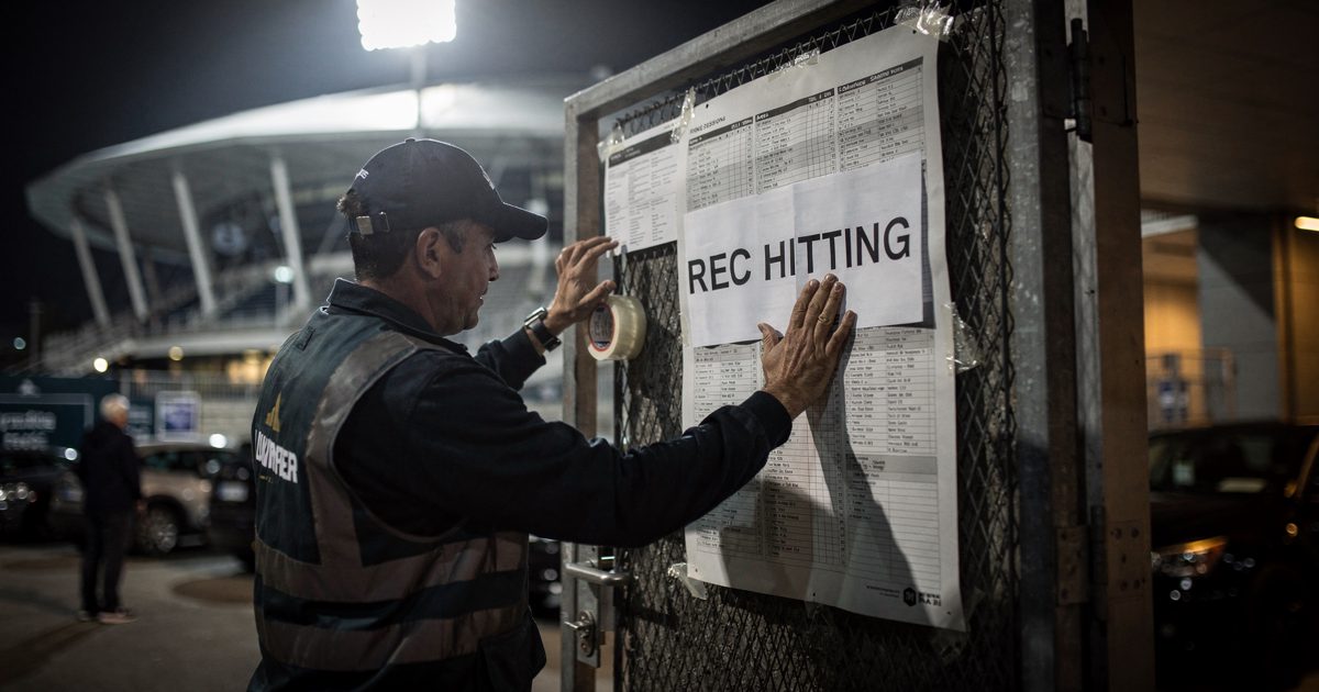 Night stadium floodlight on a service gate as a worker cropped at the hat brim presses a paper sign reading "REC HITTING" over a printed night-session draw.