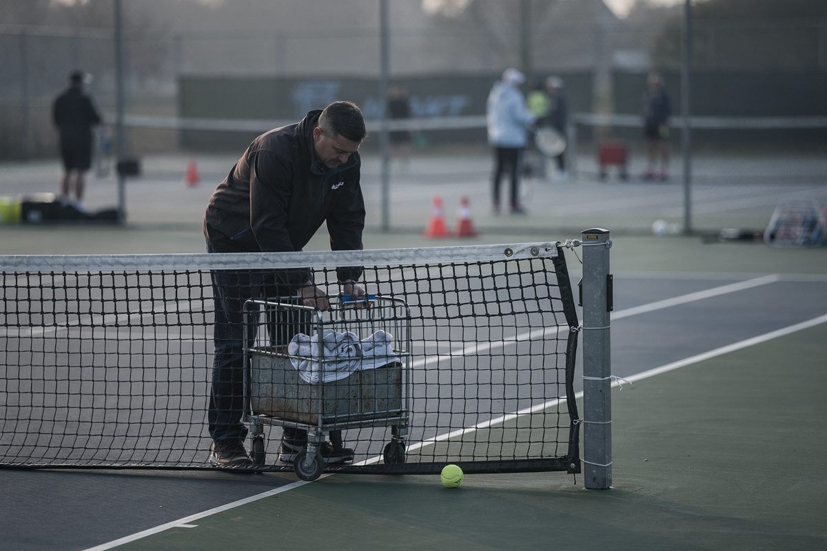Pale morning light on an empty outer tennis court as a cleaner with face turned into a collar rights a metal towel bin beside a net post, one yellow ball under.