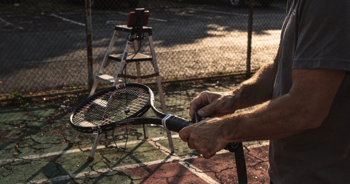 Low evening sun on a cracked public tennis court, close on a pair of hands rewrapping a racket grip with black electrical tape, a stepladder with a phone.