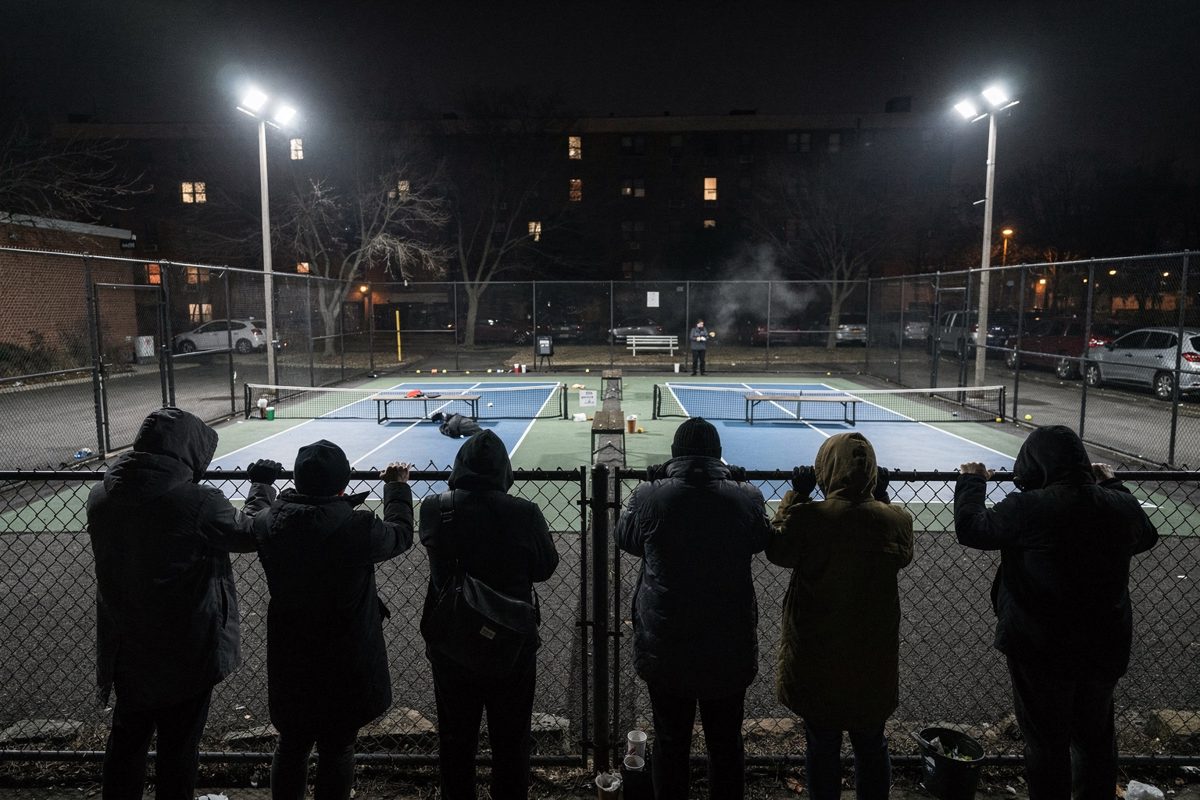 Wide night shot of three floodlit public pickleball courts behind chain-link fencing, a dozen residents in winter coats seen from behind and above pressed.