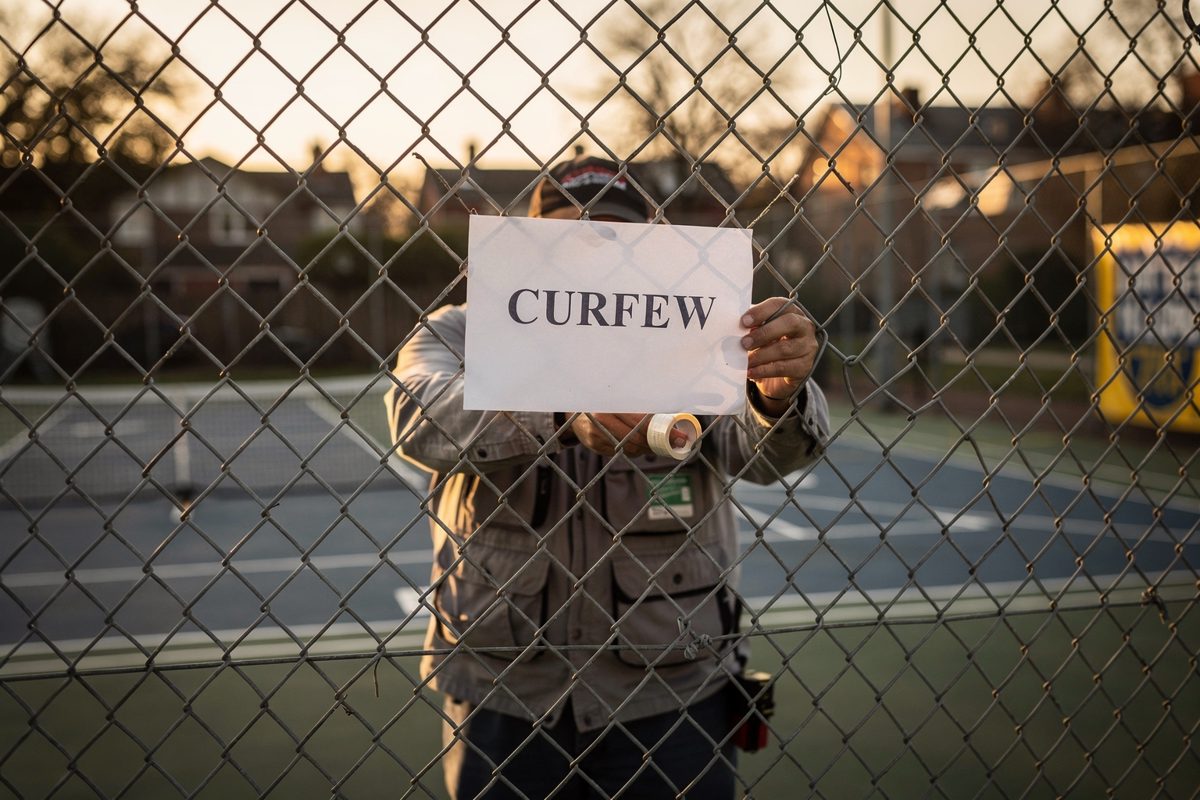Low morning sun on a chain-link fence at a municipal pickleball court, a maintenance worker cropped at the shoulders taping up a paper notice reading "CURFEW".