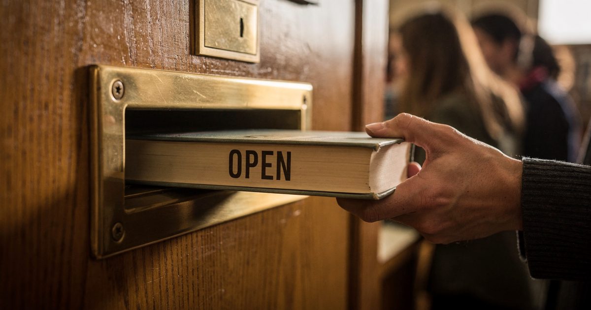 Close detail in warm late-morning light at a small public library, a hand cropped at the wrist pushing a thick hardback with a spine reading "OPEN" halfway.