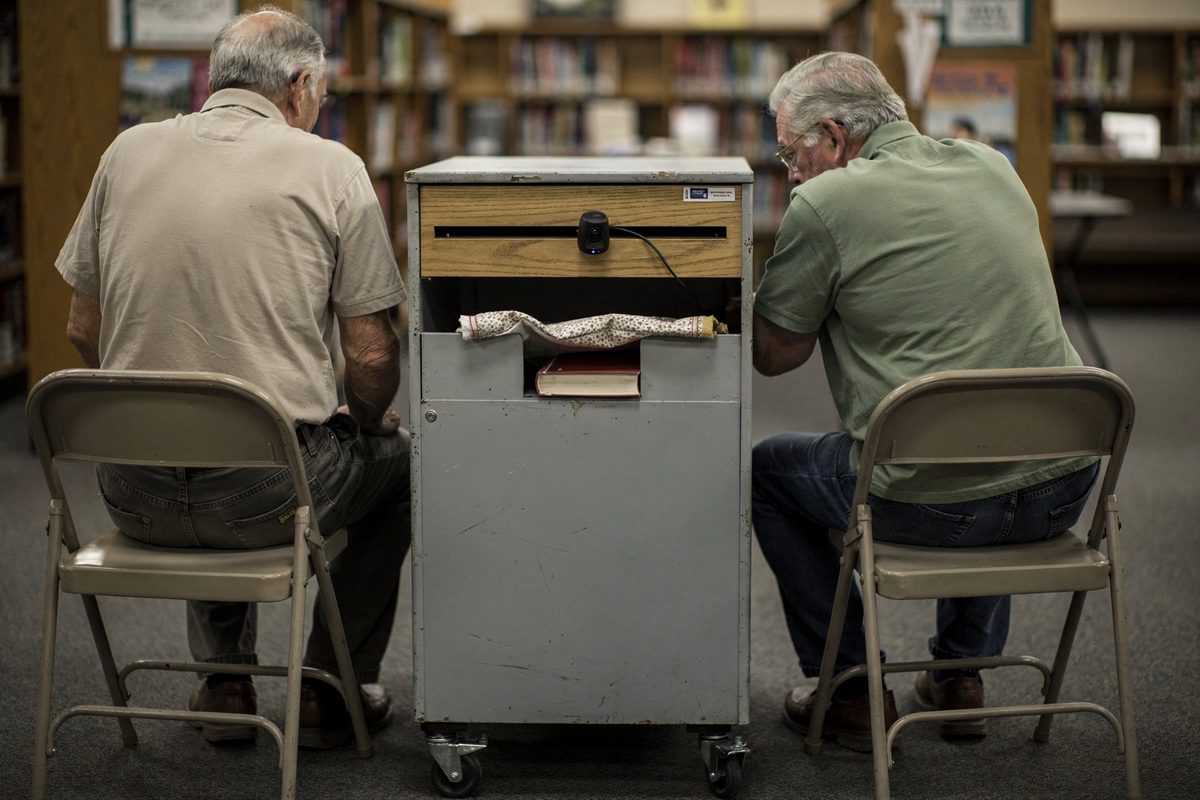 Mid-shot from behind under cool afternoon fluorescent light in a suburban branch library, two older volunteers perched on folding stools either side of a.