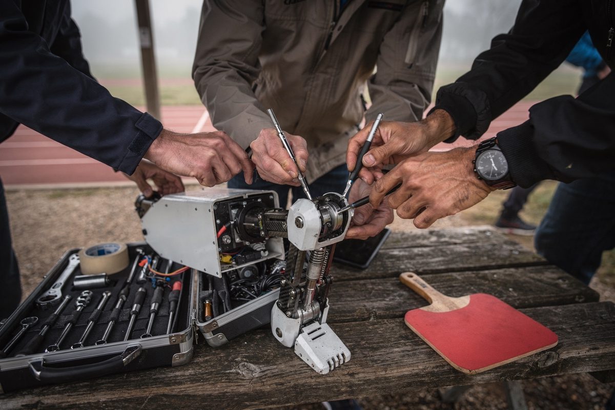 Tight shot inside a park pavilion in soft morning haze, a cluster of engineers' hands crowded around an exposed robotic knee joint, an open toolbox and a.