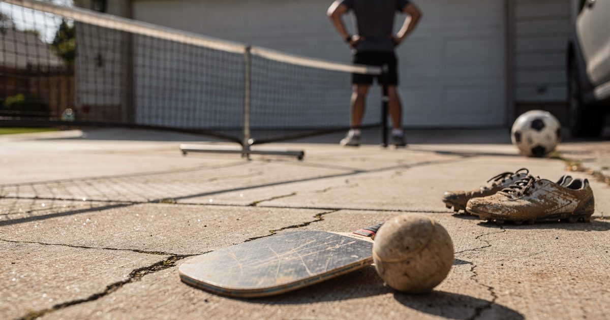 A severely cracked suburban concrete driveway featuring a sagging portable pickleball net under harsh, glaring afternoon sunlight, with a scuffed composite.