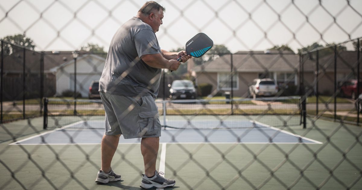 A low-angle shot peering through the rigid squares of a chain-link fence at a neighborhood pickleball court in the harsh, flat light of early Sunday morning.