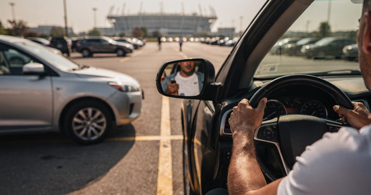 Harsh afternoon glare bakes a sprawling asphalt stadium parking lot as a driver, framed from the neck down in a white athletic top, grips a leather steering.