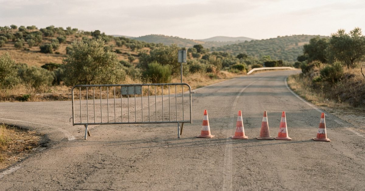 A quiet rural road in Portugal closed for a bike race: a metal crowd barrier and a line of orange traffic cones stretch across the junction, olive trees and.