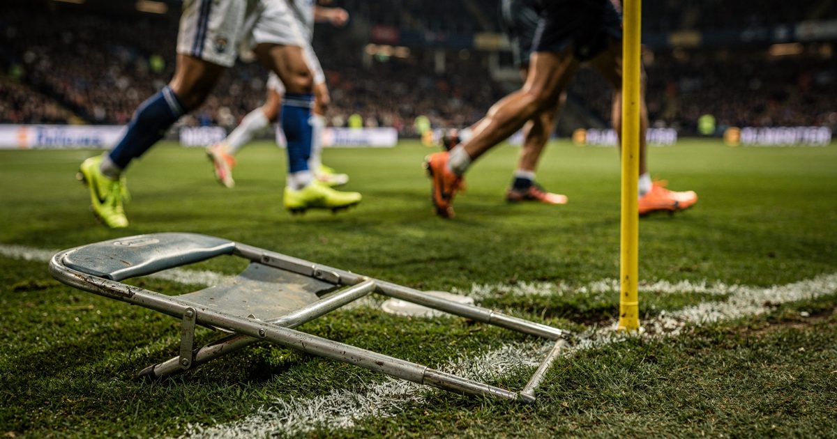 Touchline grass under bright stadium lights, a discarded steel folding chair lying on the white chalk line near a corner flag, with blurred legs in neon soccer.