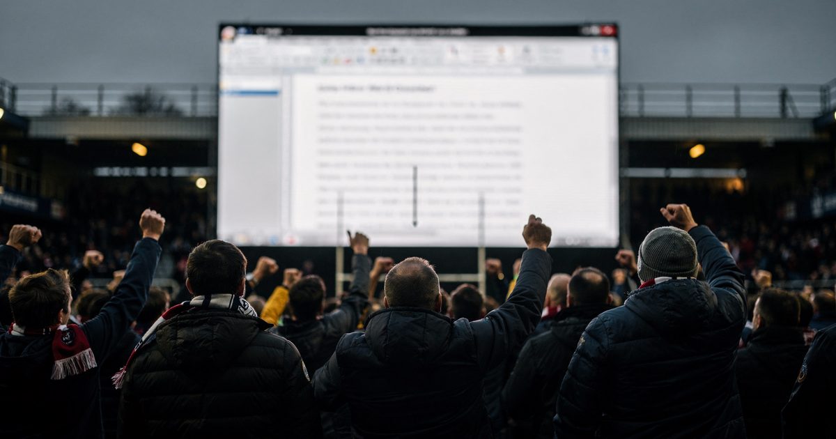 Wide shot from the back of a rugby stadium stand under evening floodlights, rows of fans seen from behind with arms and scarves raised toward a giant screen at.