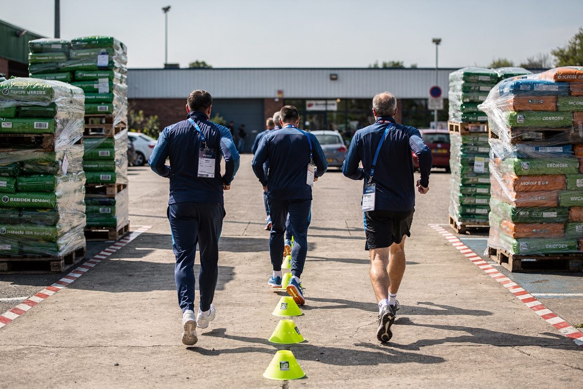 Wide shot in bright mid-morning sun across a garden centre loading bay, four men in tracksuits and lanyards seen from behind weaving a slalom between cones.