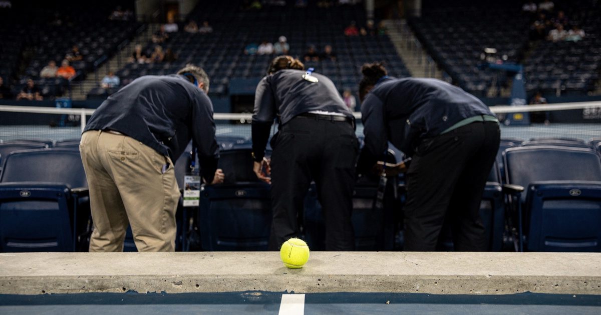 Mid-distance night view from the baseline at Arthur Ashe Stadium as three staff members with their backs to camera lean into a row of seats under white.