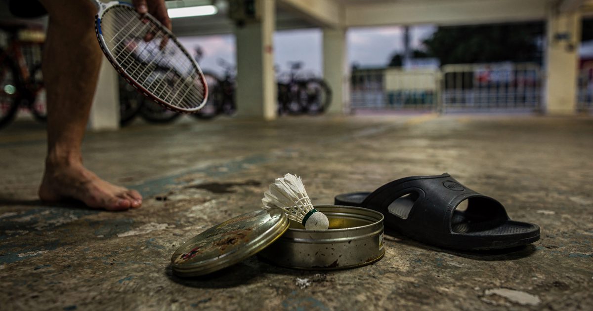 Close detail at knee height on the concrete floor of a Singapore housing-block void deck at dusk, a worn badminton shuttlecock resting inside an open round.