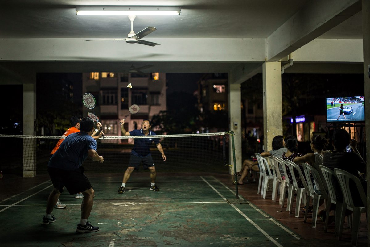Wide shot from the far end of a void deck court at night under one humming fluorescent tube, two doubles pairs seen from behind at the net with a ceiling fan.