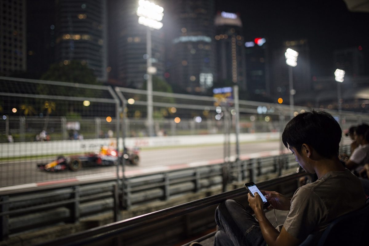 Medium shot from the grandstand side of the Marina Bay street circuit under floodlights late at night, a race car blurred past the barrier with the city.