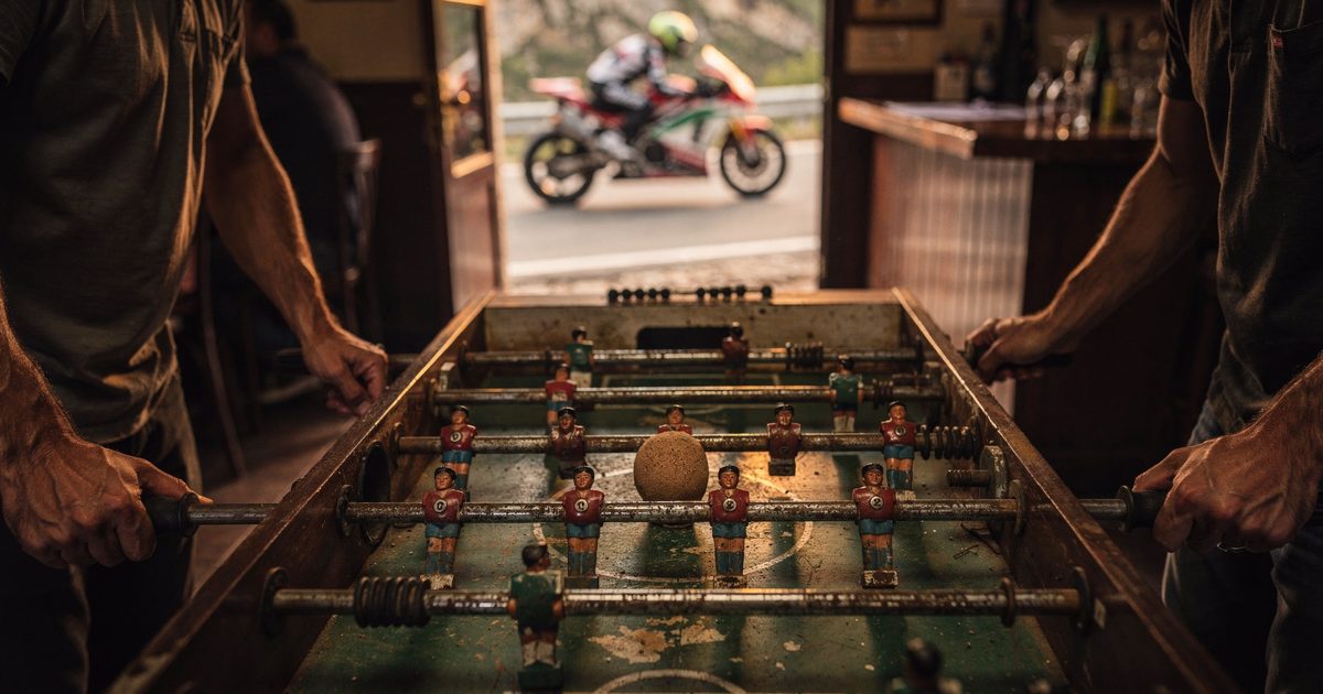Close detail in low golden late-afternoon light inside a mountain-road roadside bar, two players cropped at the forearms gripping the worn steel rods of a.