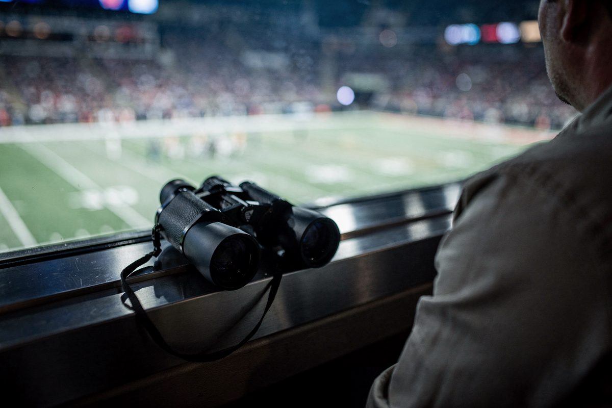 Close detail under cool blue-white stadium floodlights of unused binoculars on a suite ledge, a spectator’s shoulder cropped at the right edge and the playing.