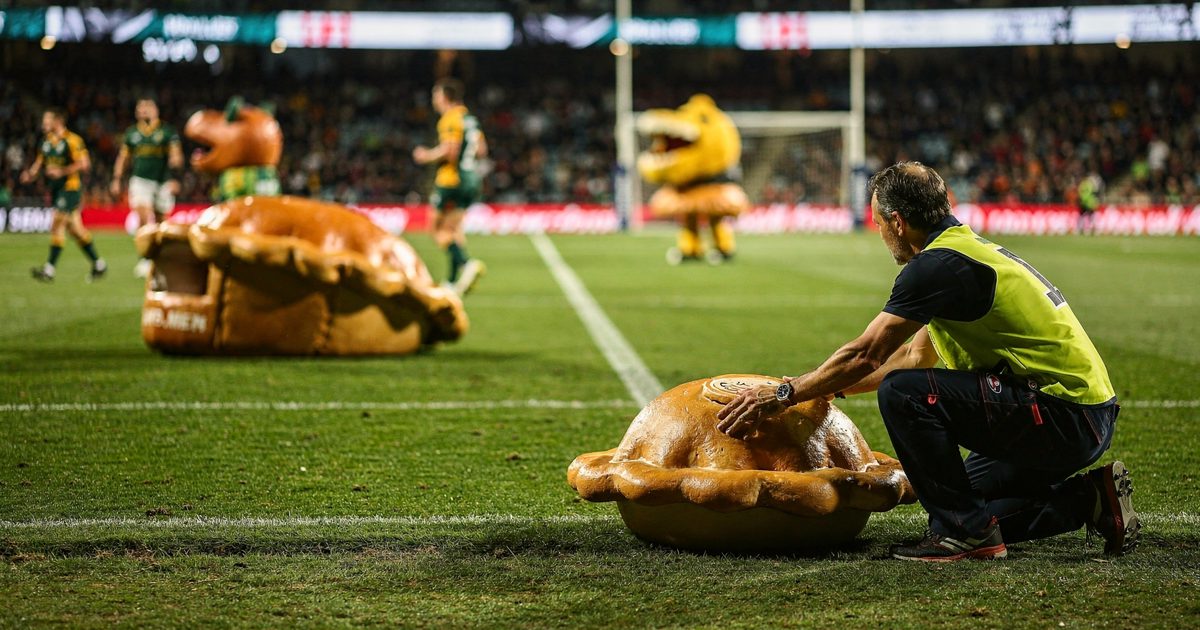 Close shot at the edge of a floodlit rugby in-goal area at night, a handler in a high-vis vest crouching to retrieve the foam lid of a giant meat pie mascot.