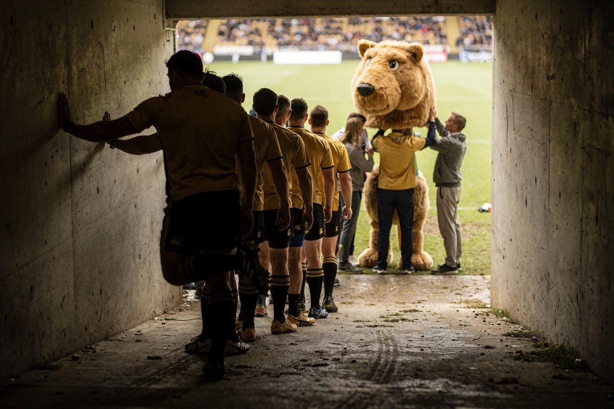 View from behind a line of rugby players in gold jerseys waiting in a concrete stadium tunnel at half-time, one stretching a calf against the wall, their eyes.
