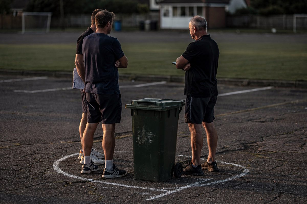 Wide evening shot from behind three men in shorts and trainers standing around a wheelie bin on cracked tarmac, arms folded, a chalk boundary line curving past.