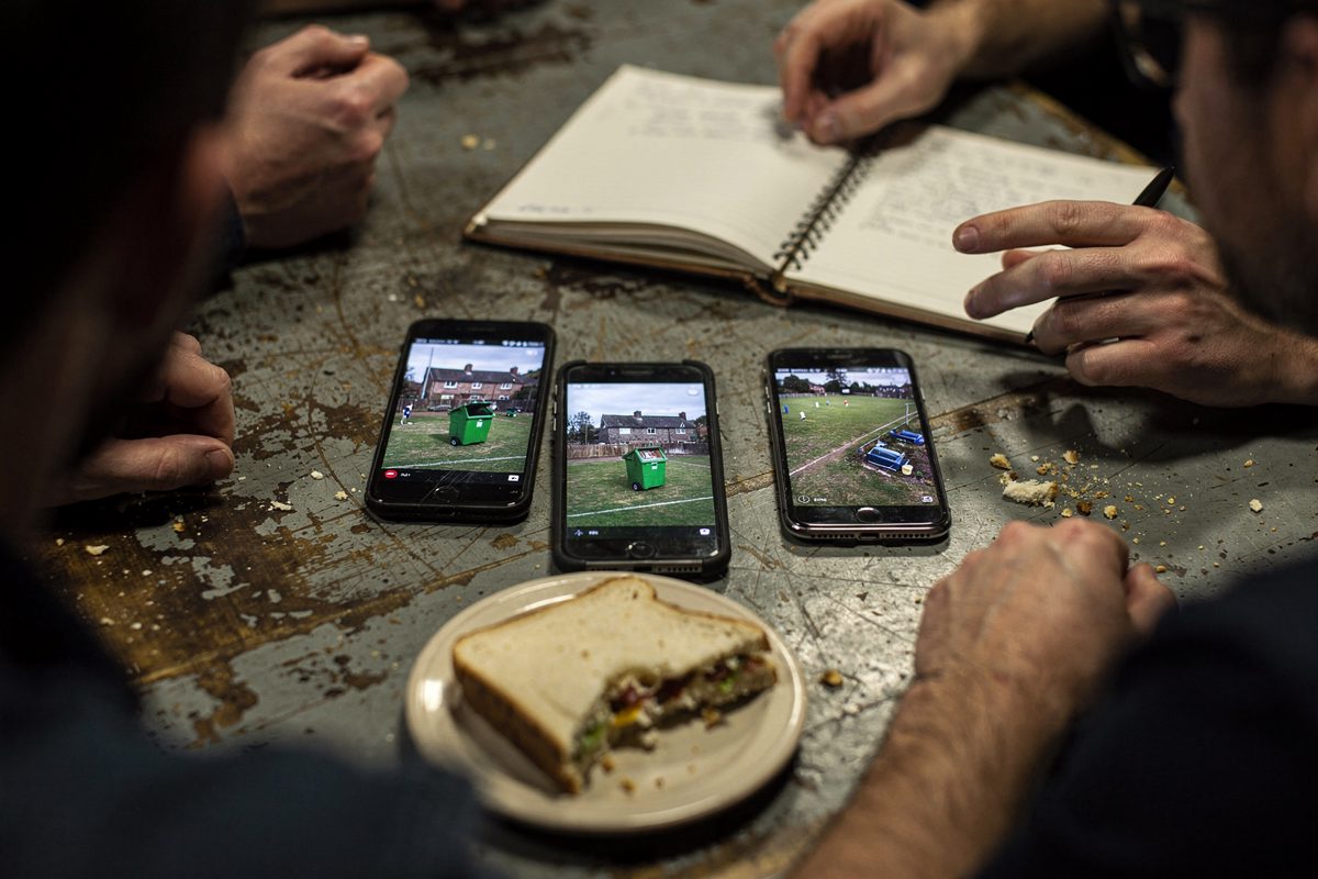 Tight overhead detail of a scuffed folding table in a pub function room under fluorescent light, three phones laid face-up beside an open spiral notebook and a.