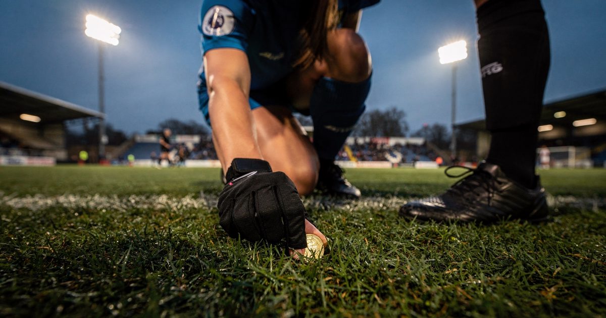 Camera lying on the turf at pitch level under white floodlights at a small south London ground at dusk, a player in a captain's armband crouched with one hand.