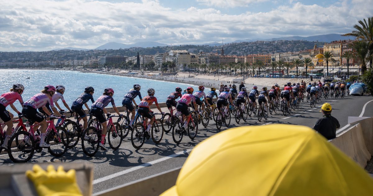 Wide editorial sports photograph of a women’s professional cycling peloton racing along a sunlit coastal road near Nice, viewed from behind a roadside barrier.