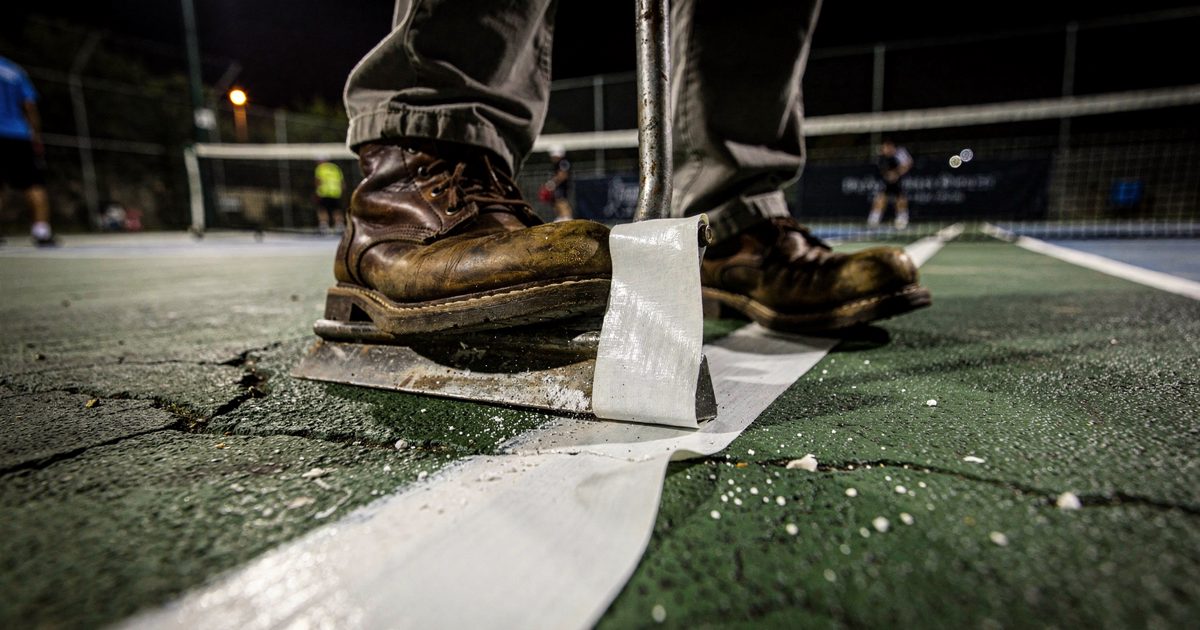 Close shot at ground level under harsh halogen floodlights on a green asphalt court, heavy leather work boots bracing as a metal floor scraper peels away the.