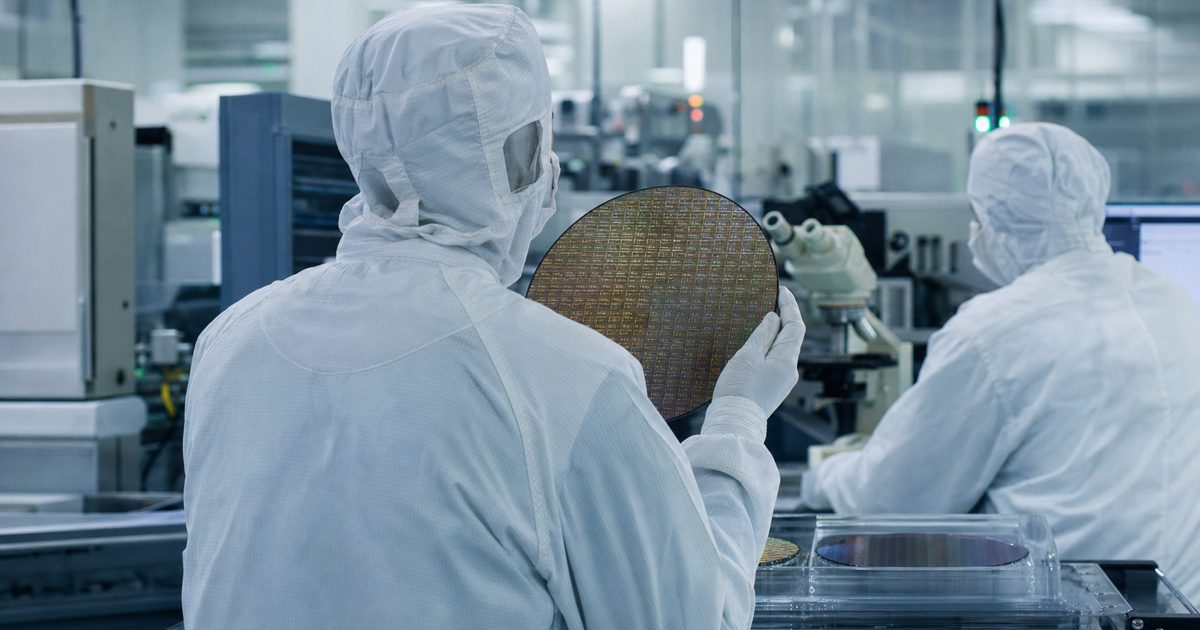 Documentary news photograph inside a modern semiconductor laboratory, showing anonymous engineers in cleanroom suits from behind inspecting silicon wafers.