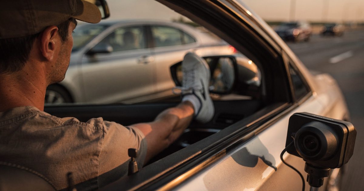 Medium shot through an open side window in hard late-afternoon sun on a multi-lane interstate, a driver visible only as a shoulder and a baseball cap, one foot.