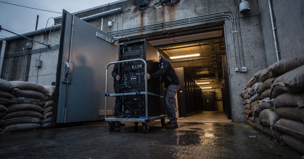 Low-angle shot down a concrete loading ramp into a data center basement at first light, two technicians seen only from the waist down guiding a tall server.