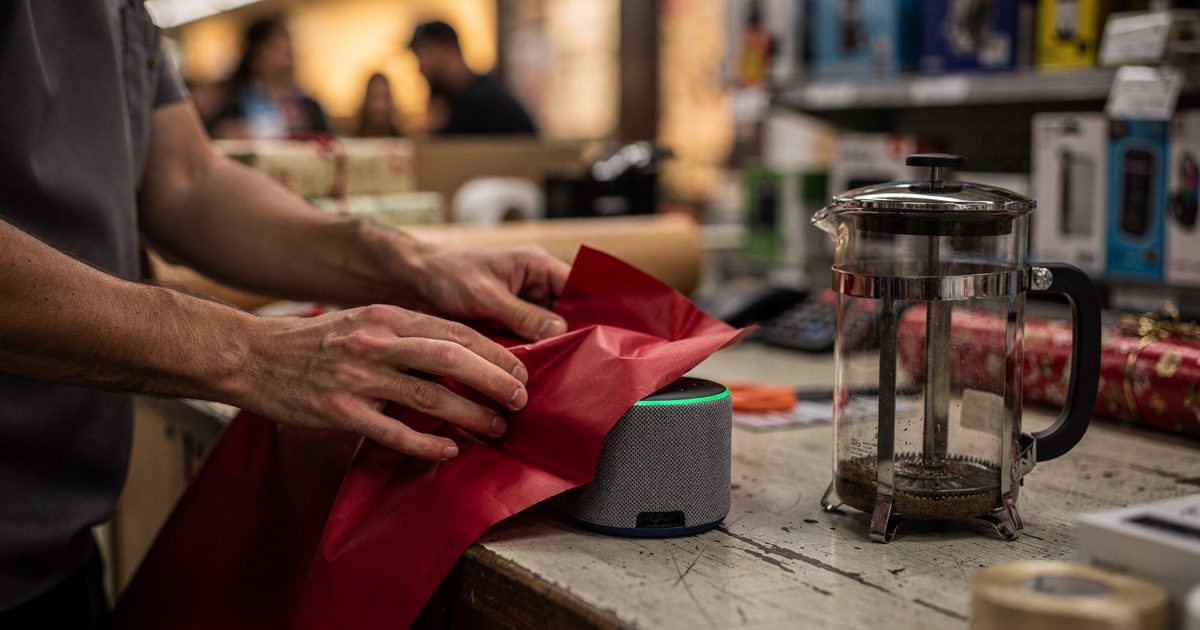 Close detail at a department store gift-wrapping counter in warm early-evening light, a clerk's hands folding red paper around a small boxed smart speaker.