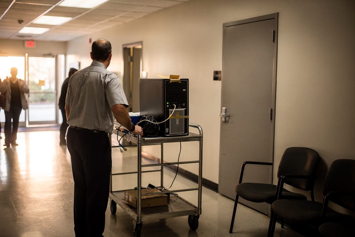 In the sunlit lobby corridor of a Montgomery county office building just after opening, a clerk photographed from behind pushes a mail cart carrying a desktop.