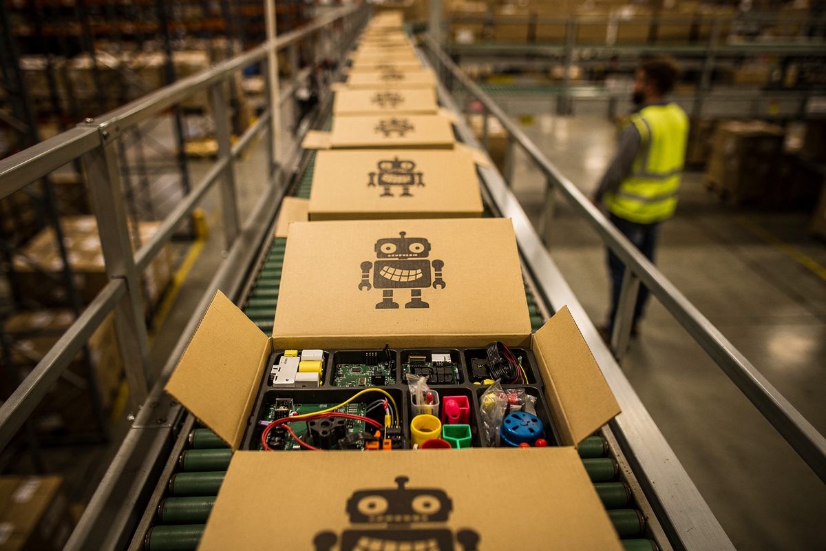 Wide shot from an overhead gantry inside a sprawling distribution warehouse under sodium lighting, a conveyor belt carrying identical cardboard boxes stamped.
