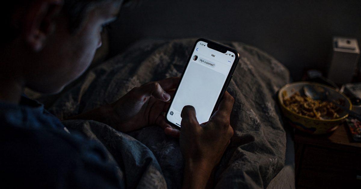 Close-up at night of a teenager's hands under a duvet holding a phone whose screen shows only a blank chat window, the glow lighting a half-eaten bowl of.
