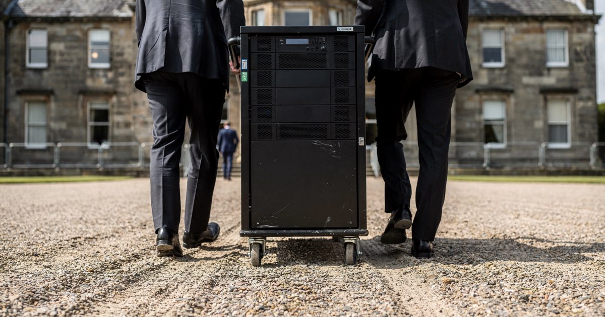 Low-angle shot in hard midday sunlight of two technicians in dark suits wheeling a tall black server cabinet across the gravel forecourt of a Scottish country.