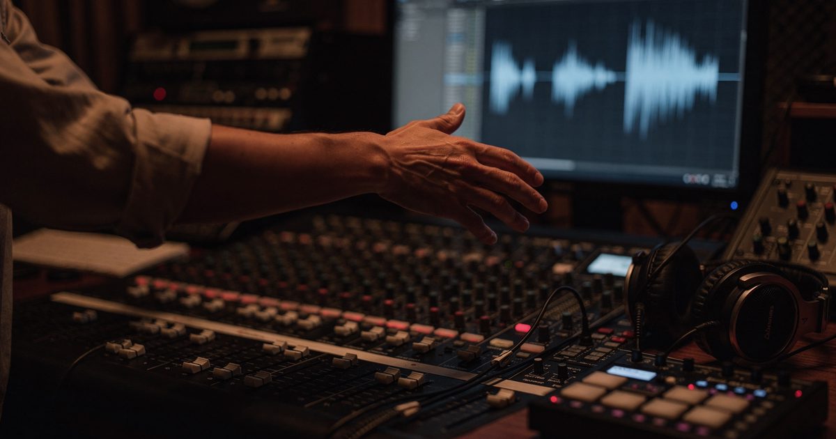 Close-up in a dim recording studio at night, a producer's hands hovering motionless above mixing-console faders while a monitor's waveform glow lights the desk.